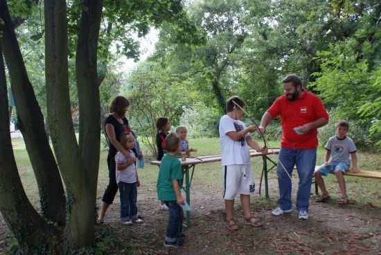 Eine Gruppe von Menschen, einschließlich Kindern, versammelt sich in einem Park. Ein Mann in einem roten T-Shirt hält einen Bogen und einen Pfeil, während andere zusehen.