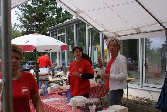 Drei Frauen in roten Shirts stehen unter einem Zelt, eine hält eine Kaffeemaschine. Ein Tisch mit Essen und Getränken ist in der Nähe aufgebaut.