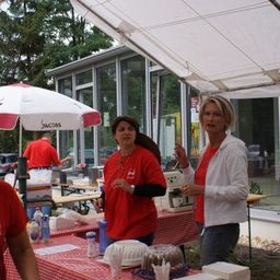 Drei Frauen in roten Shirts stehen unter einem Zelt, eine hält eine Kaffeemaschine. Ein Tisch mit Essen und Getränken ist in der Nähe aufgebaut.