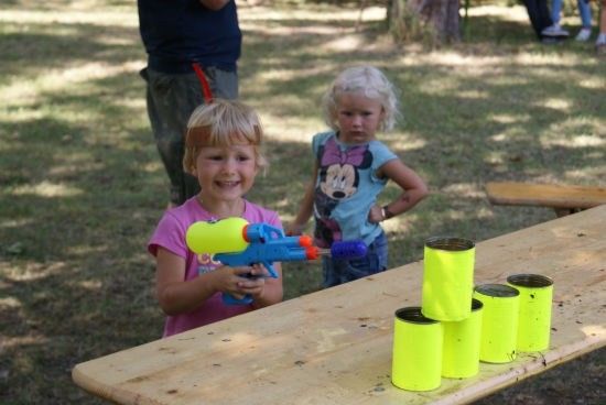 Zwei kleine Mädchen spielen mit einer Wasserpistole auf einem Holztisch mit gelben Bechern.