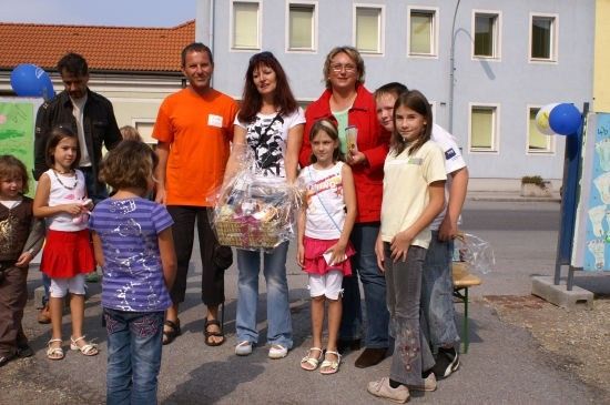 Eine Gruppe von Menschen, darunter Erwachsene und Kinder, stehen vor einem Gebäude, wobei eine Frau einen Korb mit Geschenken hält.