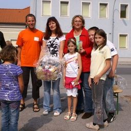 Eine Gruppe von Menschen, darunter Erwachsene und Kinder, stehen vor einem Gebäude, wobei eine Frau einen Korb mit Geschenken hält.