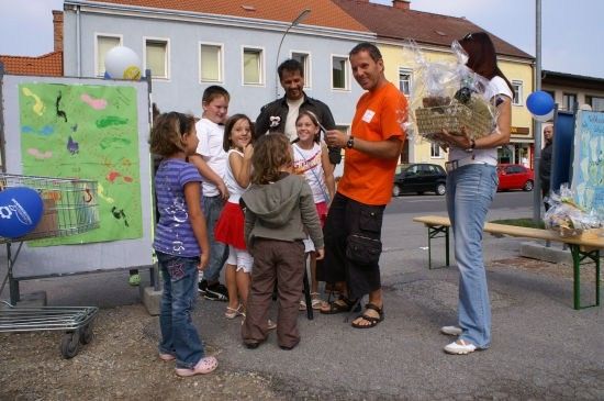 Eine Gruppe von Kindern und Erwachsenen steht vor einem Gebäude, ein Erwachsener hält einen Korb mit Essen, während ein anderer einen Ballon hält. Ein Einkaufswagen und eine Bank sind in der Nähe.