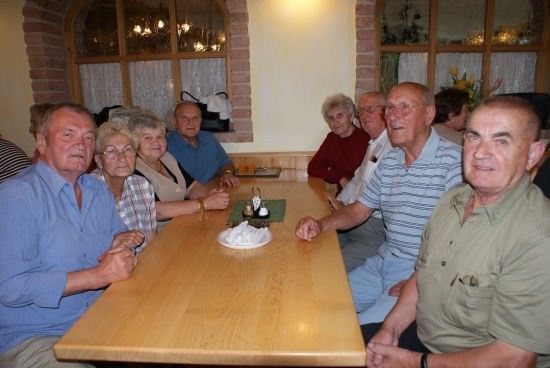 Eine Gruppe älterer Menschen sitzt um einen Holztisch in einem Restaurant. Auf dem Tisch steht ein Teller mit einem Kuchen und einer kleinen Flasche.