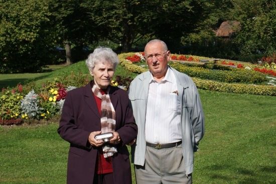Ein älteres Paar steht in einem Park, die Frau hält eine Kamera und der Mann trägt eine Brille. Sie sind von einem farbenfrohen Blumenbeet und grünem Rasen umgeben.