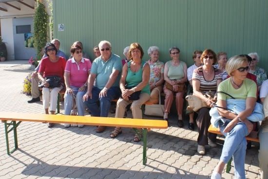 Eine Gruppe von Menschen, überwiegend Frauen, sitzt auf Bänken vor einem grünen Gebäude. Sie sind lässig gekleidet und scheinen auf etwas zu warten.