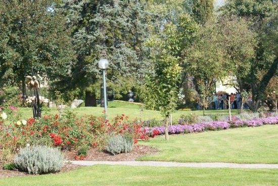 Ein gepflegter Park mit einer Vielzahl bunter Blumen, saftigem grünem Gras und Bäumen. Ein Laternenpfahl steht in der Mitte, und eine Gruppe von Menschen ist in der Nähe versammelt.