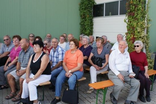 Eine Gruppe älterer Menschen sitzt draußen auf Bänken, einige mit Brille und Sonnenbrille, vor einer grünen Wand mit Pflanzen.