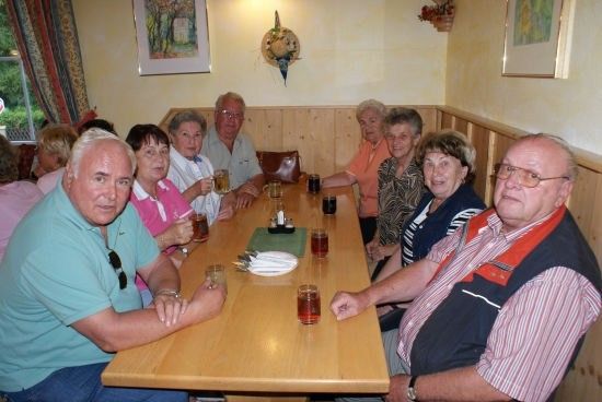 Eine Gruppe älterer Menschen sitzt in einem Restaurant um einen Holztisch herum und hält Gläser mit Getränken in den Händen. Sie lächeln und scheinen ihre Mahlzeit zu genießen.