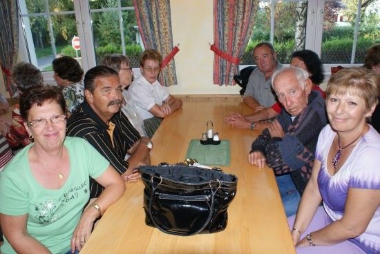 Eine Gruppe von Menschen sitzt um einen Tisch mit einer schwarzen Tasche darauf. Sie sind in einem Raum mit großen Fenstern und Vorhängen.
