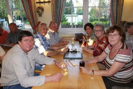 Eine Gruppe älterer Menschen sitzt um einen Tisch herum, hält Gläser mit Getränken in der Hand und posiert für ein Foto. Der Tisch ist mit verschiedenen Gegenständen gedeckt, und der Raum hat große Fenster mit Vorhängen.