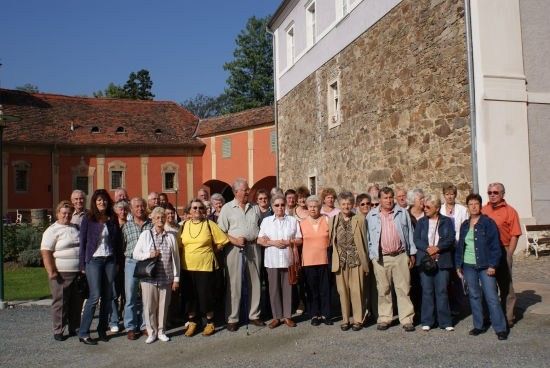 Eine Gruppe von Menschen, hauptsächlich ältere, posiert für ein Foto vor einem Steingebäude. Das Gebäude hat ein rotes Dach und weiße Wände. Der Himmel ist klar und blau.