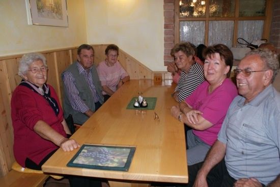 Eine Gruppe älterer Menschen sitzt um einen Holztisch in einem gemütlichen Raum, lächelt und schaut in die Kamera.