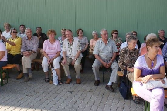 Eine Gruppe älterer Menschen sitzt auf Bänken vor einer grünen Wand, einige tragen Sonnenbrillen und Halsketten.