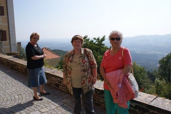 Drei Frauen stehen auf einem Kopfsteinpflasterweg und lächeln für ein Foto mit einer malerischen Aussicht auf Berge und Grünflächen im Hintergrund.