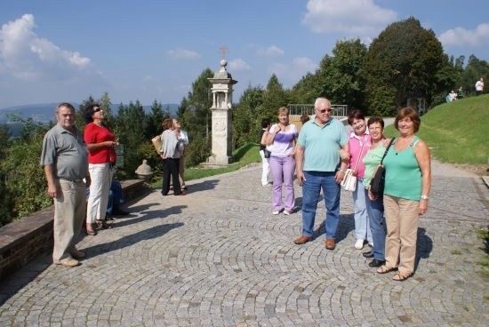 Eine Gruppe von Menschen, einschließlich Erwachsener, steht auf einem Kopfsteinpflasterweg, möglicherweise auf einem Hügel, mit einem Steindenkmal und Bäumen im Hintergrund.