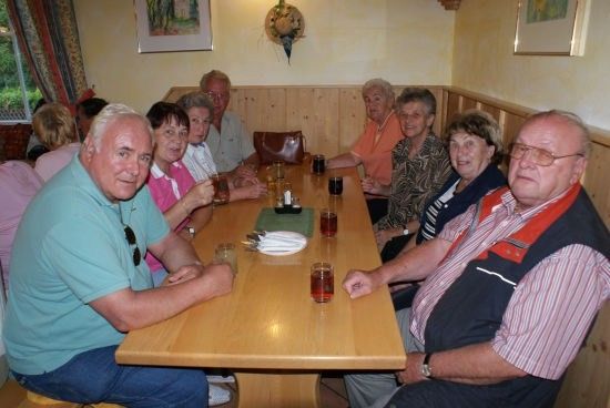 Eine Gruppe älterer Menschen sitzt um einen Holztisch in einem Restaurant, lächelt und hält Gläser mit Getränken in den Händen.