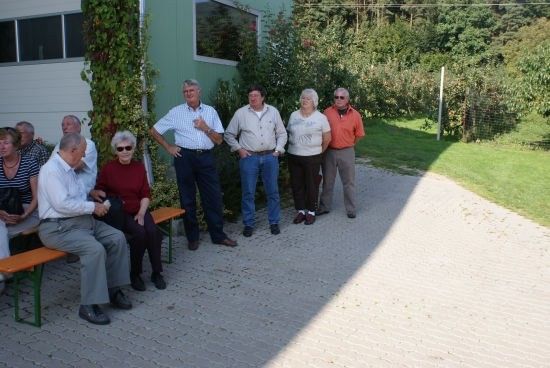 Eine Gruppe von Menschen, einige stehend und einige auf Bänken sitzend, versammelt sich vor einem Gebäude mit Grünpflanzen im Hintergrund.