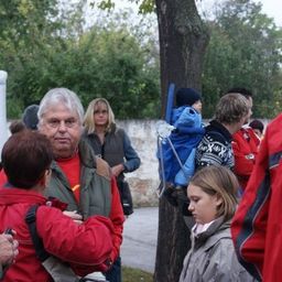 Eine Gruppe von Menschen in roten Jacken versammelt sich um einen Baum, ein Mann in einem roten Shirt und eine Frau mit einem Baby im Kinderwagen.