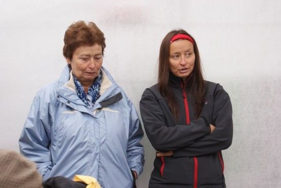 Zwei Frauen stehen nebeneinander, eine mit einer blauen Jacke und die andere in einer schwarzen Jacke mit einem roten Stirnband, beide schauen nach vorne.