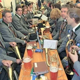 Eine Gruppe von Musikern in Uniform sitzt an einem langen Tisch mit verschiedenen Musikinstrumenten und Getränken, möglicherweise in Vorbereitung auf eine Aufführung.
