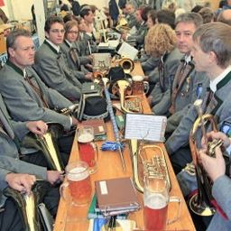 Eine Gruppe von Musikern in Uniform sitzt an einem langen Tisch mit verschiedenen Musikinstrumenten und Getränken, möglicherweise in Vorbereitung auf eine Aufführung.