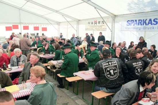 Eine große Gruppe von Menschen in grünen Uniformen und Hüten sitzt unter einem Zelt an Tischen, einige spielen Musikinstrumente. Es gibt Schilder am Zelt, eines heißt 'HAMER' und ein anderes 'SPARKAS'.