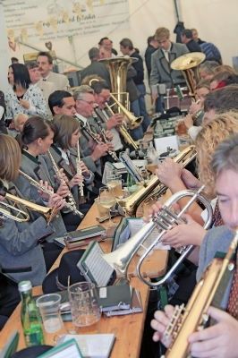Eine Gruppe von Musikern spielt verschiedene Blechblasinstrumente, darunter Trompeten und Tuben, bei einer Veranstaltung. Einige sitzen, andere stehen, alle konzentrieren sich auf ihre Instrumente. Der Ort scheint eine große Zusammenkunft zu sein, möglicherweise ein Konzert oder Festival.