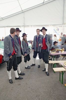 Fünf Personen in traditionellen Uniformen, einschließlich Hüten und Stiefeln, stehen in einer Reihe bei einer Veranstaltung im Freien. Hinter ihnen ist ein Tisch mit einem Musikinstrument und einer Bank zu sehen, und ein weißes Zelt ist im Hintergrund.