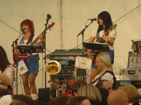 Zwei Frauen spielen Gitarren auf der Bühne bei einem Konzert, mit einem Posaune und Keyboard dahinter. Ein Publikum beobachtet von vorne.