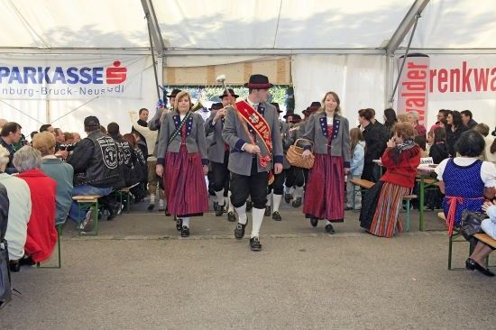 Eine Gruppe von Menschen in traditionellen Kostümen, die in einer Reihe unter einem Zelt mit einem Banner, das 'Parkasse' steht, gehen. Sie sind von einem Publikum umgeben, das auf Bänken sitzt.
