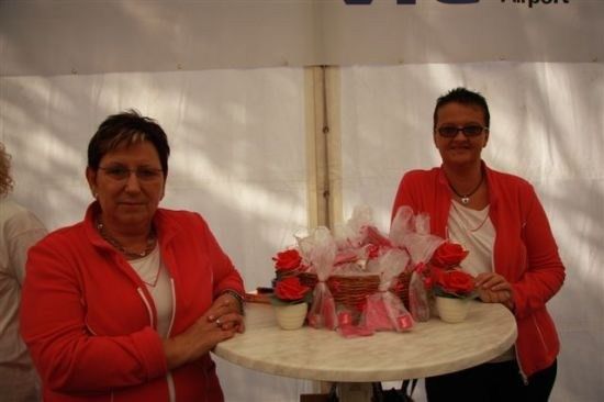 Zwei Frauen in roten Jacken stehen neben einem Tisch mit Blumenarrangements und lächeln für ein Foto.