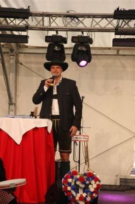 Ein Mann in traditioneller bayerischer Kleidung steht auf der Bühne, hält ein Glas Bier in der Hand, mit einem roten Tischtuch und einem Kranz hinter ihm.