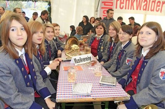 Eine Gruppe junger Frauen in einheitlichen Outfits sitzt um einen Tisch mit verschiedenen Gegenständen, darunter ein Schild mit der Aufschrift 'Musikvereine'. Sie scheinen sich bei einer Veranstaltung zu befinden.
