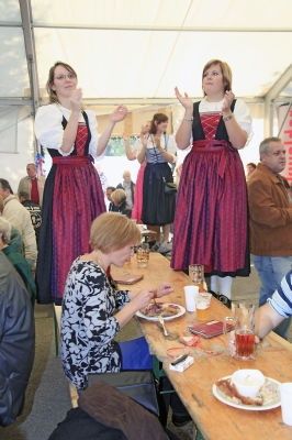 Drei Frauen in traditioneller Kleidung führen auf der Bühne eines Festivals auf, während eine sitzende Frau vor einem langen Tisch eine Mahlzeit genießt.