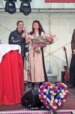 Ein Mann und eine Frau stehen auf einer Bühne mit einem Blumenstrauß, einer roten Tischdecke und einem herzförmigen Blumenarrangement im Vordergrund. Im Hintergrund ist eine Gitarre zu sehen.
