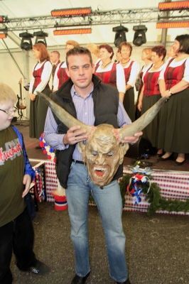 Ein Mann hält eine gehörnte Maske vor einer Bühne, auf der eine Gruppe von Frauen in traditioneller Kleidung singt.