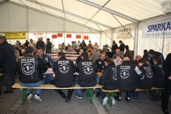 Eine Gruppe von Menschen in passenden Jacken sitzt auf Bänken unter einem Zelt, einige mit Tellern Essen. Das Zelt hat Banner und Flaggen, und mehr Menschen sind im Hintergrund versammelt.