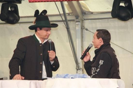 Zwei Männer sitzen an einem Tisch, einer trägt einen schwarzen Hut und der andere hält ein Mikrofon. Sie scheinen in ein Gespräch vertieft zu sein.
