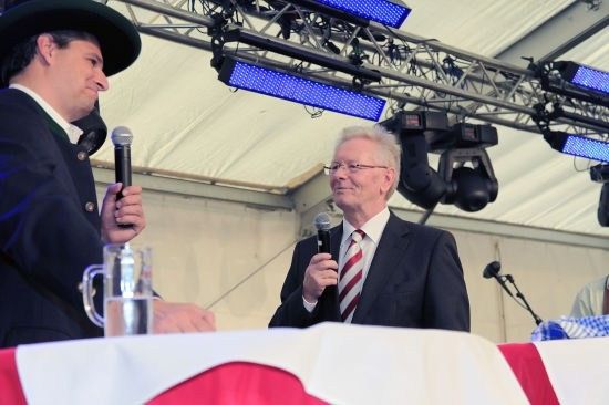 Zwei Männer in Anzügen, einer mit einem Mikrofon, stehen hinter einem Tisch mit Flaggenhintergrund, wahrscheinlich auf einer Bühne.
