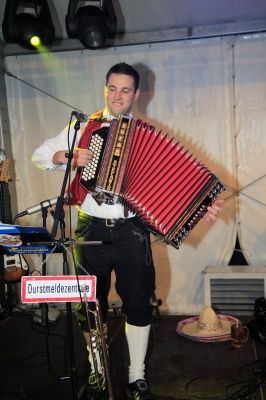 Ein Mann spielt Akkordeon auf der Bühne vor einem Mikrofon. Er trägt ein traditionelles Outfit und einen Hut. Hinter ihm steht ein Schild, das 'Durstmeldezentrum' besagt.