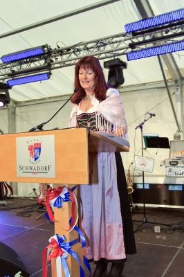 Eine Frau steht an einem Podium mit Mikrofon, in traditioneller Kleidung. Hinter ihr ist eine Bühne mit Musikinstrumenten und Lautsprechern aufgebaut. Das Podium hat ein Schild mit der Aufschrift 'Schwadorf'.