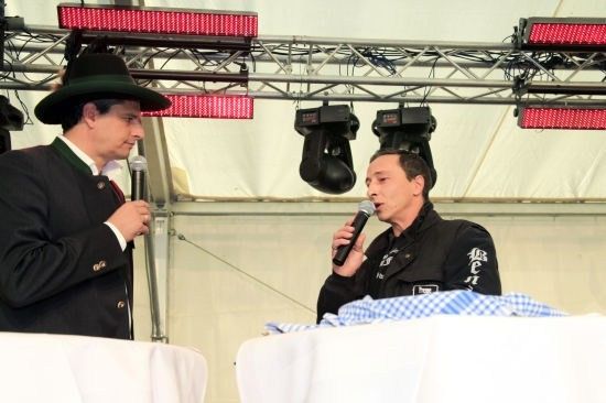 Zwei Männer in Anzügen und Hüten sprechen in Mikrofone auf einer Bühne mit roten Lichtern darüber.