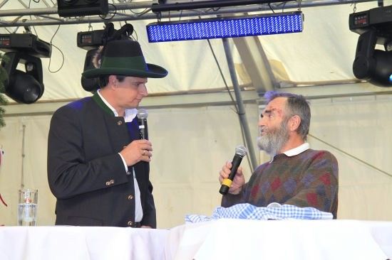 Zwei Männer sitzen an einem Tisch, einer trägt einen Hut und hält ein Mikrofon, der andere mit Bart hält ebenfalls ein Mikrofon. Sie führen wahrscheinlich ein Gespräch.