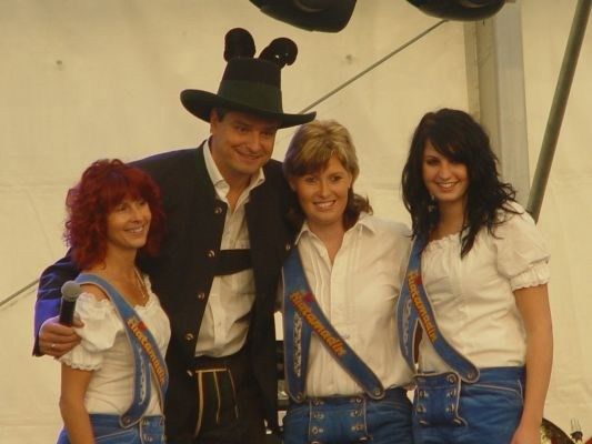 Ein Mann und drei Frauen in traditioneller bayerischer Kleidung posieren für ein Foto. Der Mann trägt einen schwarzen Hut mit Ohren und hält ein Mikrofon. Die Frauen tragen weiße Blusen mit blauen Schürzen und Hosenträger. Sie alle lächeln und stehen eng beieinander.
