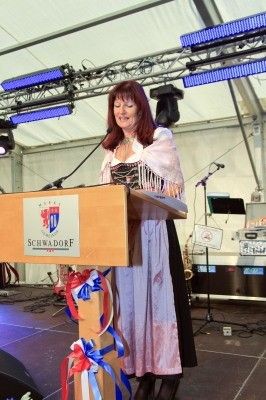 Eine Frau in einem traditionellen Kleid steht an einem Rednerpult mit einem Mikrofon, möglicherweise hält sie eine Rede. Das Rednerpult ist mit Bändern und einem Schild, das 'Schwadorf' besagt, dekoriert. Hinter ihr befindet sich eine Bühneneinrichtung mit Lautsprechern, einem Saxophon und anderen Musikinstrumenten.