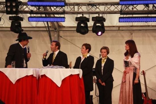 Eine Gruppe von Menschen steht auf einer Bühne, einer hält ein Mikrofon. Sie sind in formeller Kleidung gekleidet, und vor ihnen stehen Tische mit Mikrofonen.