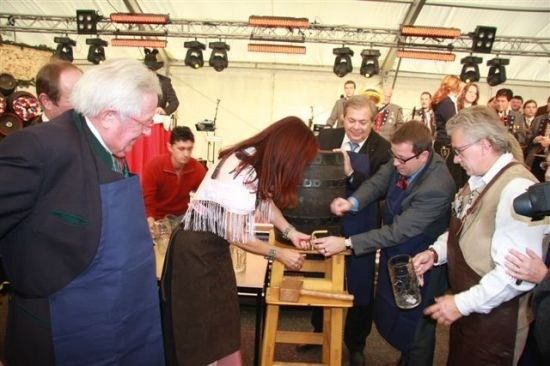 Eine Gruppe von Menschen, darunter Männer in Anzügen und eine Frau in traditioneller Kleidung, versammelt sich um einen großen Holzfass, möglicherweise zur Vorbereitung einer zeremoniellen Veranstaltung.