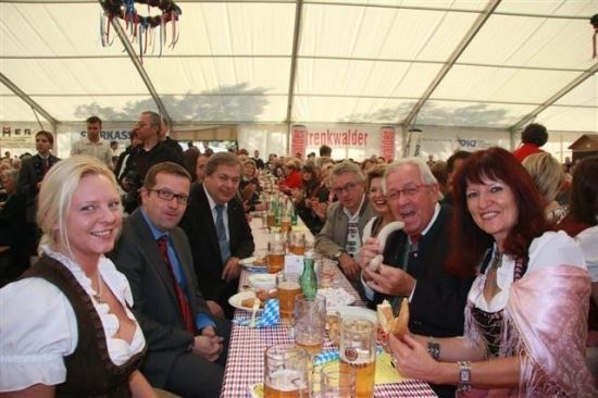 Eine Gruppe von Menschen, darunter Männer in Anzügen und Frauen in traditioneller Kleidung, sitzen an einem langen Tisch in einem Zelt und genießen Essen und Getränke. Der Tisch ist mit einer rot-weiß karierten Tischdecke bedeckt.