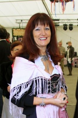 Eine Frau mit roten Haaren, die einen rosafarbenen, mit Fransen besetzten Schal und ein schwarz-weißes Oberteil trägt, lächelt in die Kamera. Sie steht in einem überfüllten Innenbereich, mit anderen Menschen im Hintergrund.